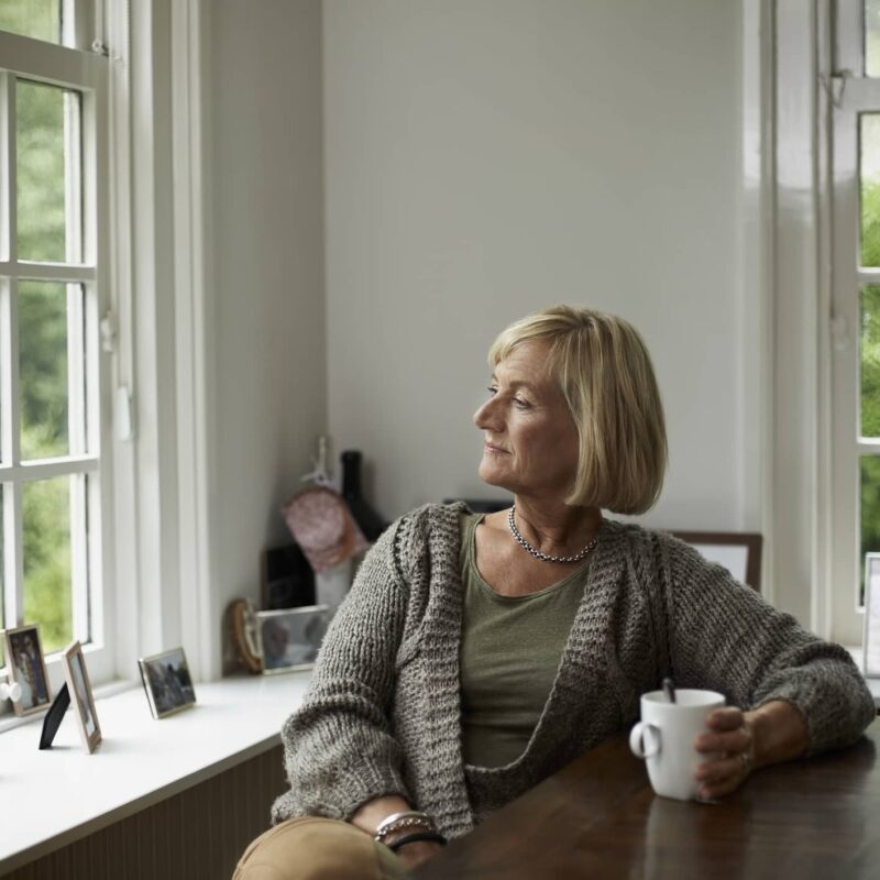 Vrouw in een woonkamer die naar buiten kijkt en haar koffie opdrinkt.